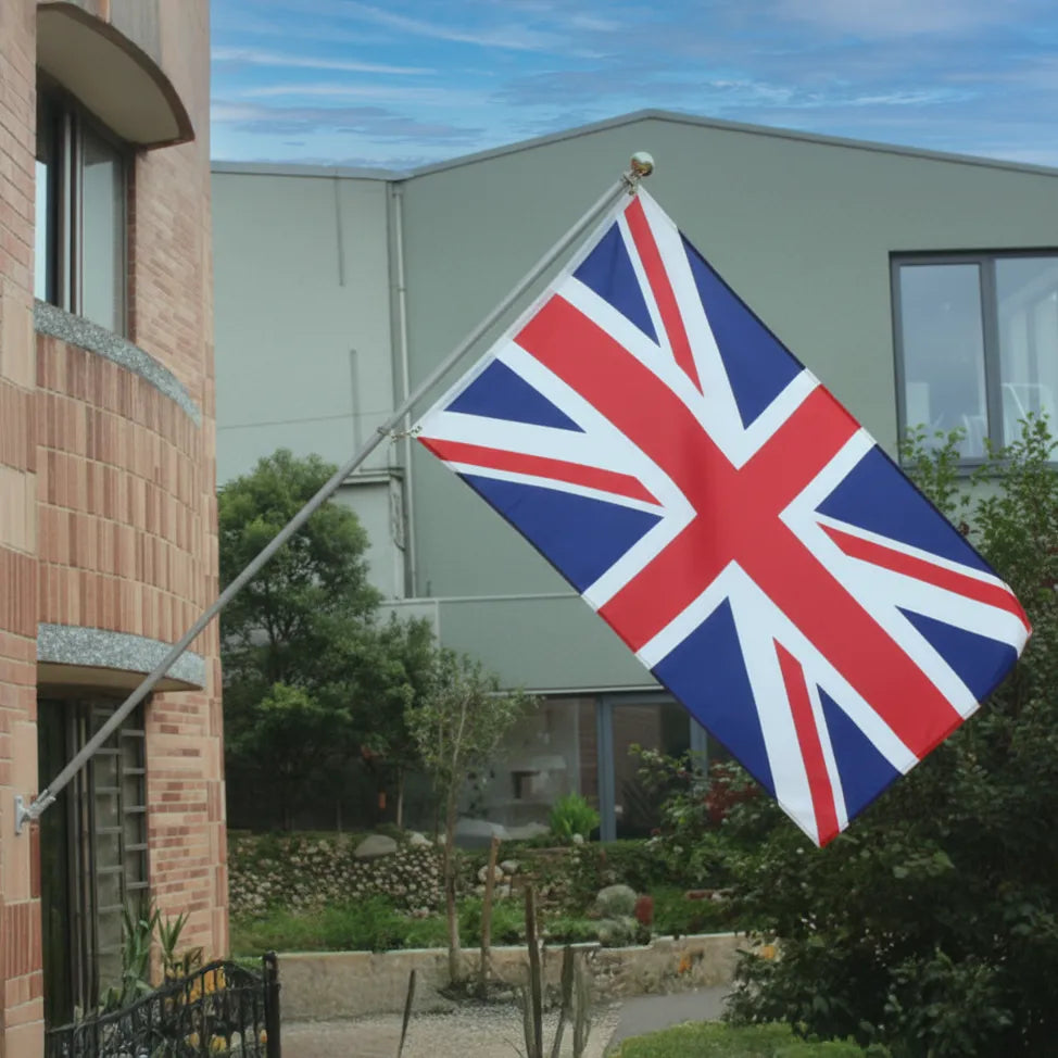 Union Jack flag waving on a lightweight wall mounted flagpole in front of a building with a garden in the background